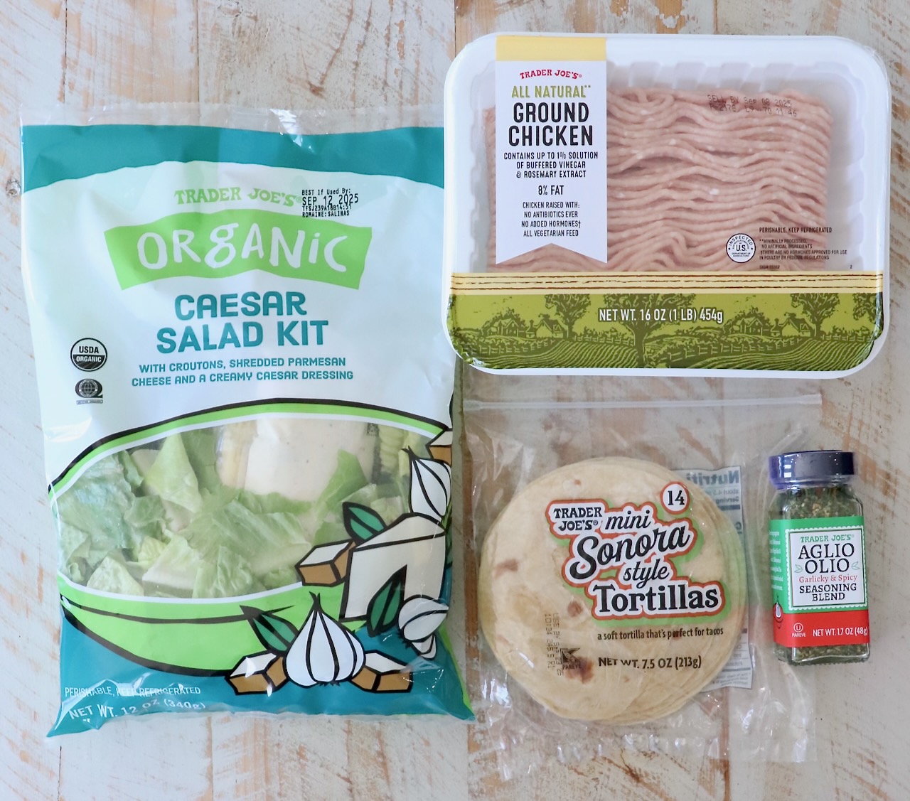 Trader Joe’s products on a wooden surface: organic Caesar salad kit, ground chicken, mini Sonora tortillas, and Aglio Olio seasoning blend.