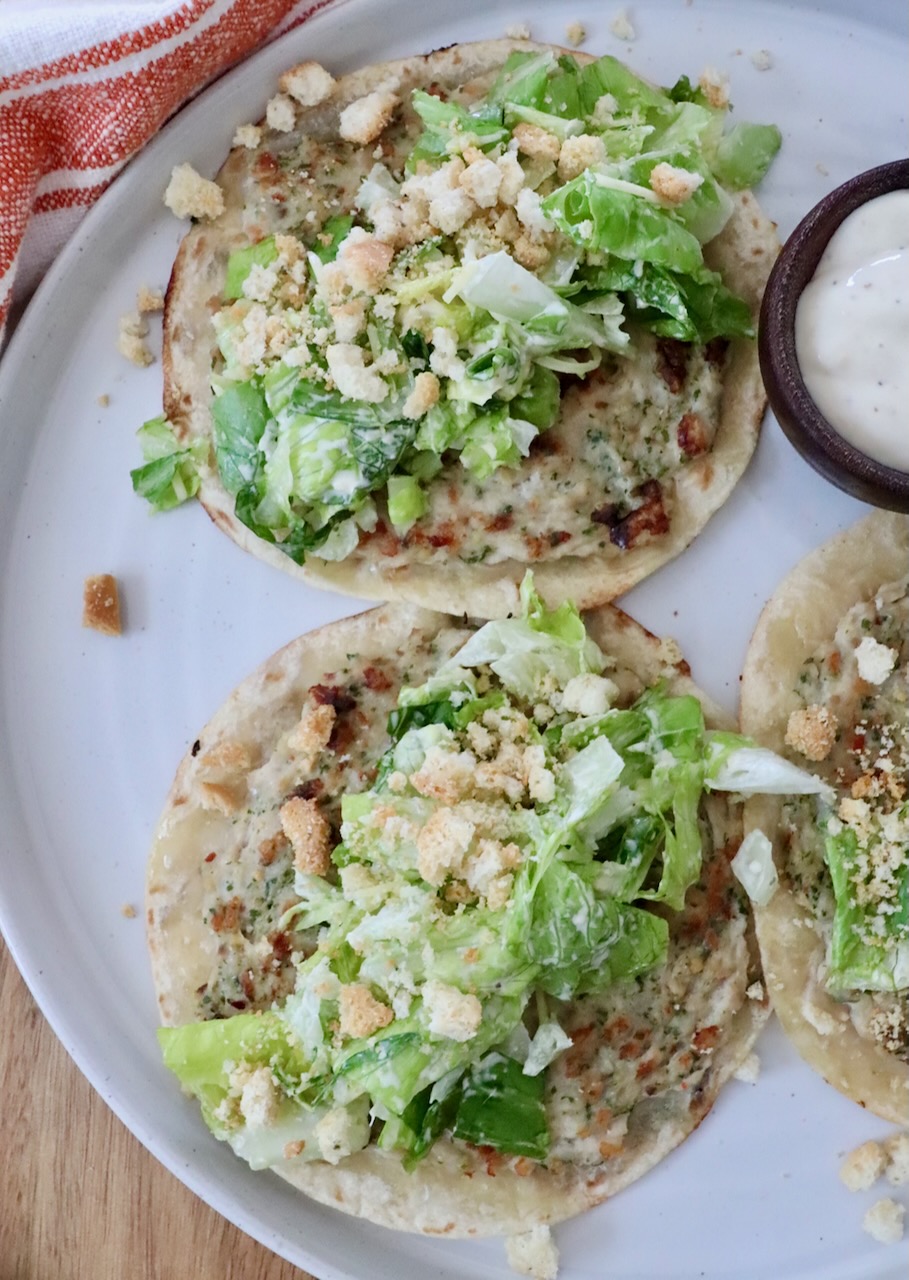Three mini Caesar salad tacos topped with lettuce, croutons, and creamy dressing on a white plate. A nearby small bowl contains additional dressing.
