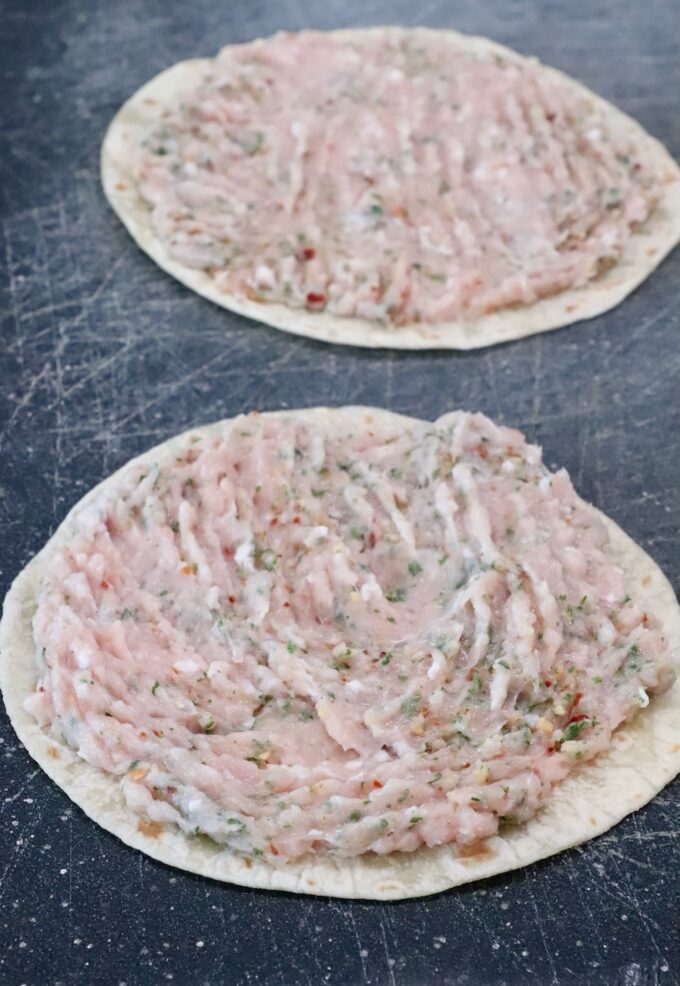 Two tortillas on a dark cutting board, each topped with a spread of raw ground chicken mixed with herbs and spices
