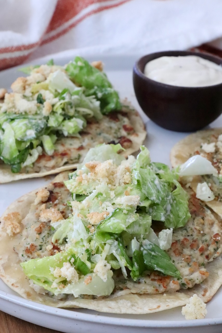 Two smash chicken tacos topped with leafy green salad and crumbled croutons on a white plate. A small bowl of creamy dipping sauce is on the side.
