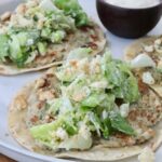 Two smash chicken tacos topped with leafy green salad and crumbled croutons on a white plate. A small bowl of creamy dipping sauce is on the side.
