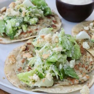 Two smash chicken tacos topped with leafy green salad and crumbled croutons on a white plate. A small bowl of creamy dipping sauce is on the side.