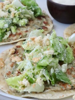 Two smash chicken tacos topped with leafy green salad and crumbled croutons on a white plate. A small bowl of creamy dipping sauce is on the side.