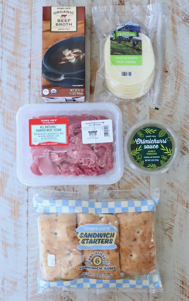 Various Trader Joe's products displayed on a wooden surface, including beef broth, provolone cheese, shaved beef steak, chimichurri sauce, and sandwich buns.