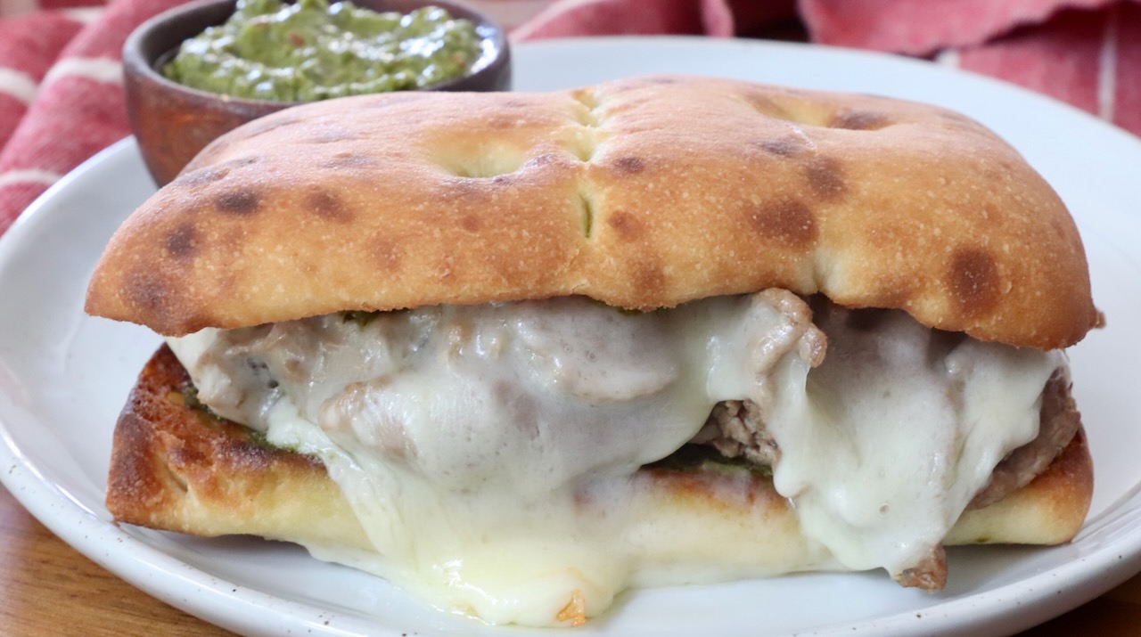 A toasted sandwich with melted cheese and meat is on a white plate. A small bowl of green chimichurri sauce is in the background, with a red cloth nearby.