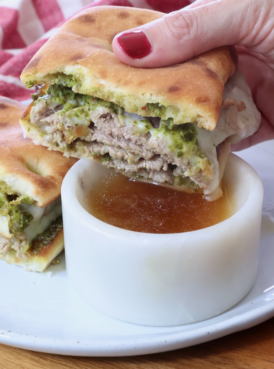 A hand with red nail polish holds a sliced sandwich filled with green chimichurri sauce and cooked steak above a small white bowl of beef broth on a plate.