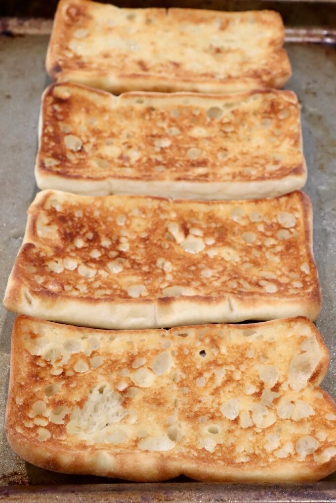 Golden-brown grilled sandwich bread slices on a baking tray, with a crispy, textured surface. 