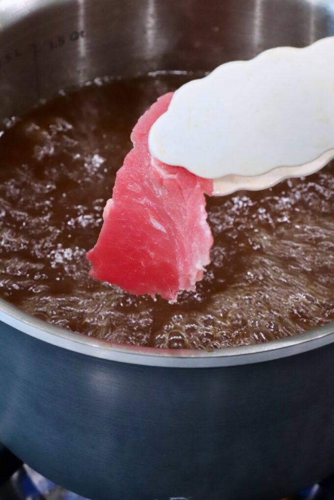 A slice of raw red meat held by tongs is about to be dipped into a pot of bubbling brown broth
