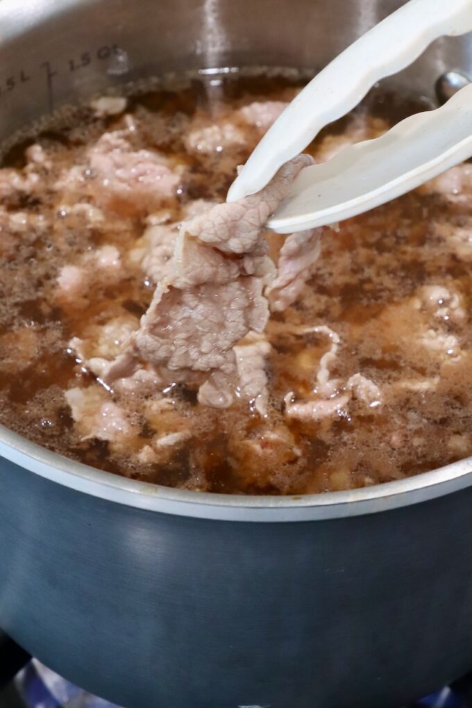A pot of brown broth with simmering beef slices is being cooked. A pair of tongs holds a piece of cooked beef above the liquid