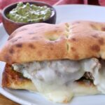 A toasted sandwich with melted cheese and meat is on a white plate. A small bowl of green chimichurri sauce is in the background, with a red cloth nearby.