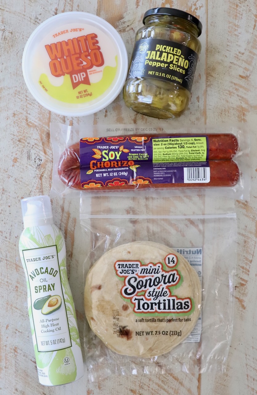 A flat lay of Trader Joe's products on a wooden surface, including white queso dip, pickled jalapeños, soy chorizo, mini tortillas, and avocado oil spray.