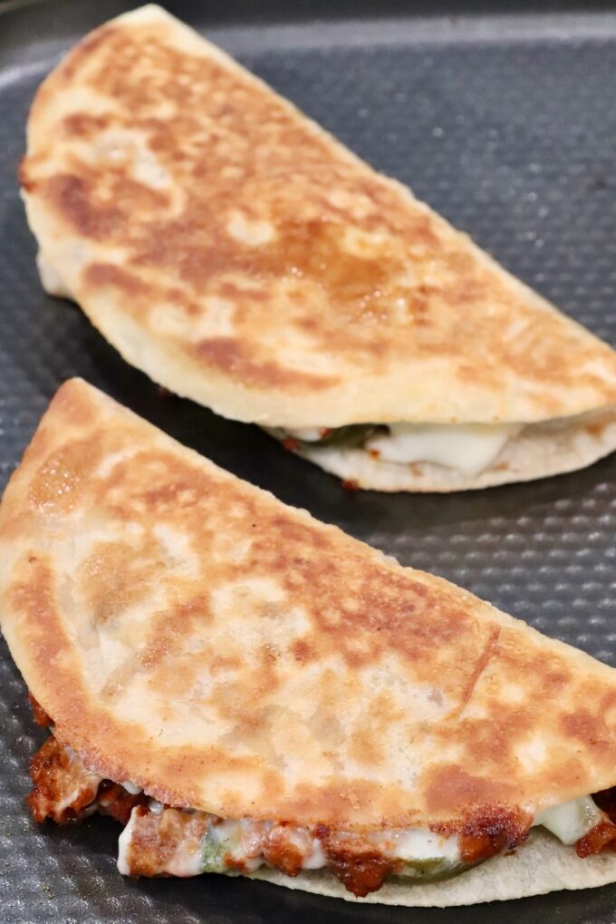 Two golden-brown crispy tacos sit in a pan, filled with melted cheese and chorizo.
