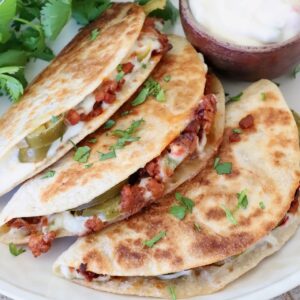 Three crispy tacos filled with chorizo, queso dip, and jalapeños are garnished with cilantro. A small bowl of white queso sits beside them on a plate.