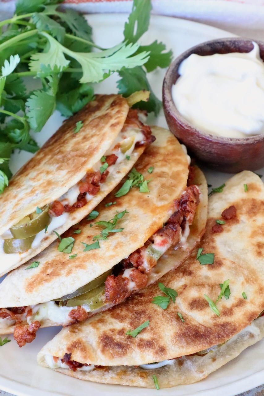 Three crispy tacos filled with meat, cheese, and jalapeños are garnished with cilantro. A small bowl of white queso sits beside them.