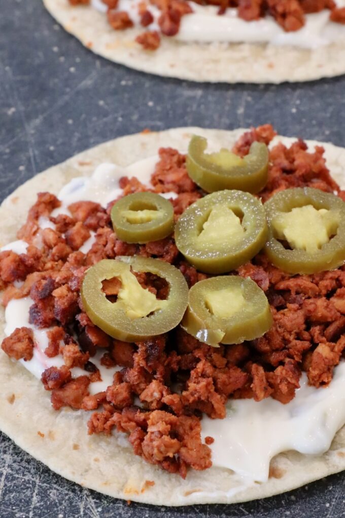 Close-up of a tortilla topped with creamy queso dip, crumbled chorizo, and sliced jalapeños on a dark surface