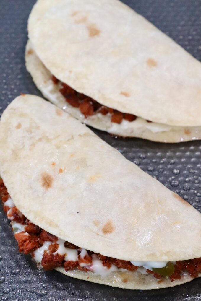 Two folded soft tacos filled with chorizo and melted cheese on a textured black surface. 