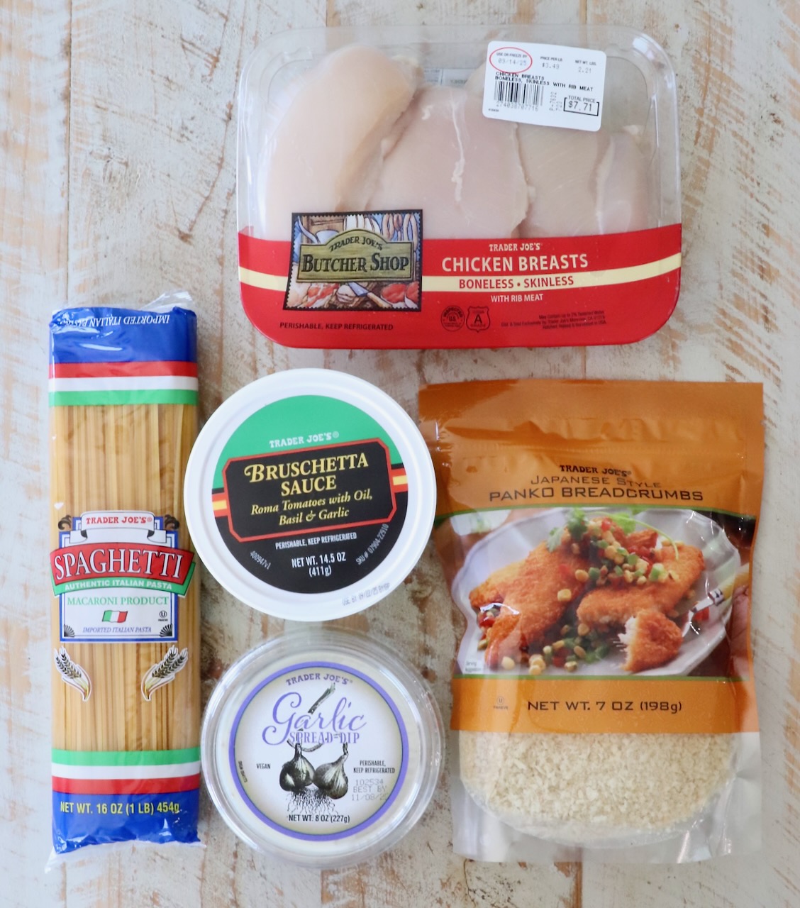 A selection of Trader Joe's products on a light wooden surface, including chicken breasts, bruschetta sauce, spaghetti, garlic spread, and panko breadcrumbs.