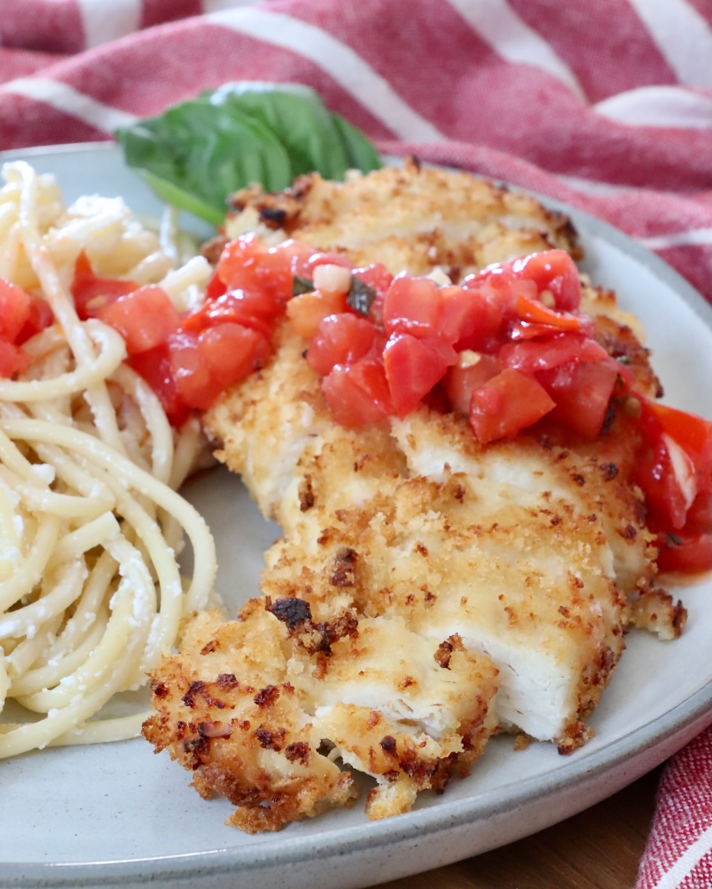 sliced piece of crispy chicken on a plate with pasta, topped with tomato bruschetta