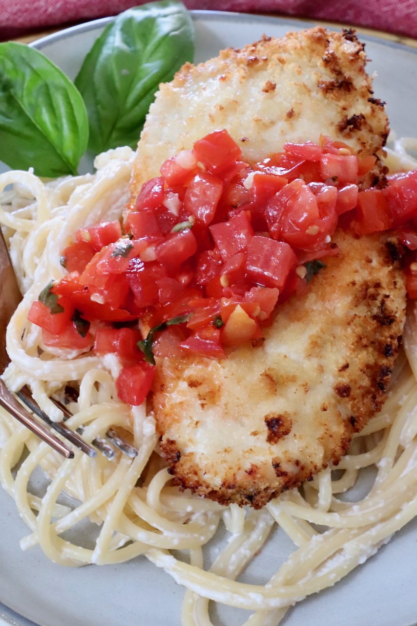 piece of crispy chicken on top of pasta on a plate, topped with tomato bruschetta, with basil leaves in the background