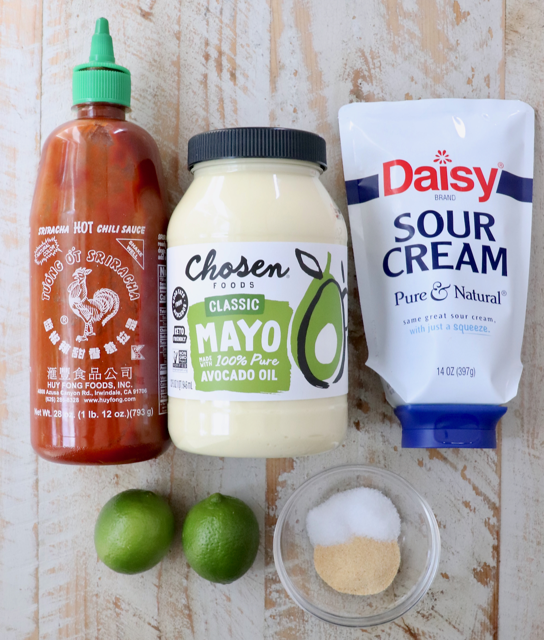 Condiments and ingredients on a wooden surface: a bottle of Sriracha hot sauce, avocado oil mayo, sour cream, two limes, and a small bowl of salt and garlic powder.