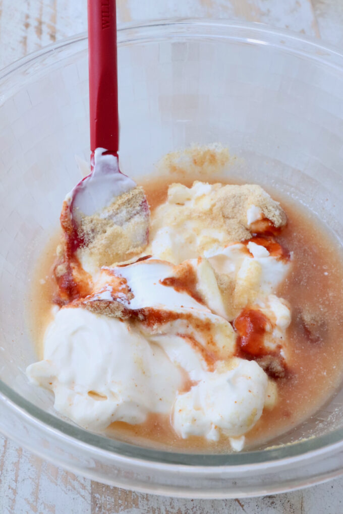Mixing bowl with white yogurt, hot sauce, and spices, partially mixed with a red spatula.