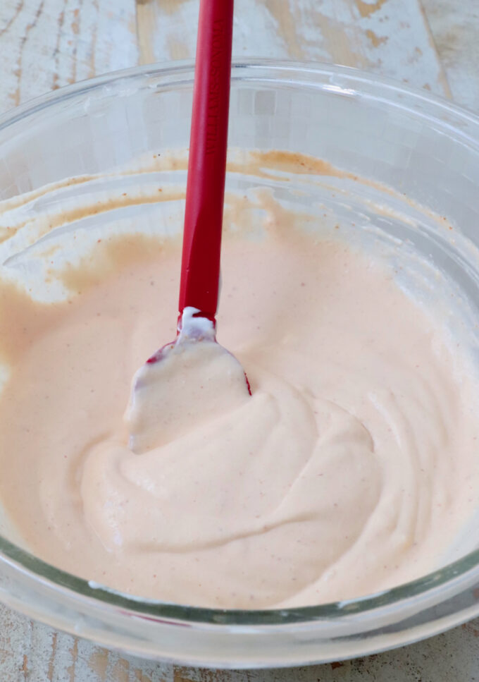A clear glass bowl filled with creamy orange sauce, being mixed with a red spatula.
