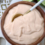 A wooden bowl filled with creamy, light orange sauce. A spoon dips into the smooth mixture.