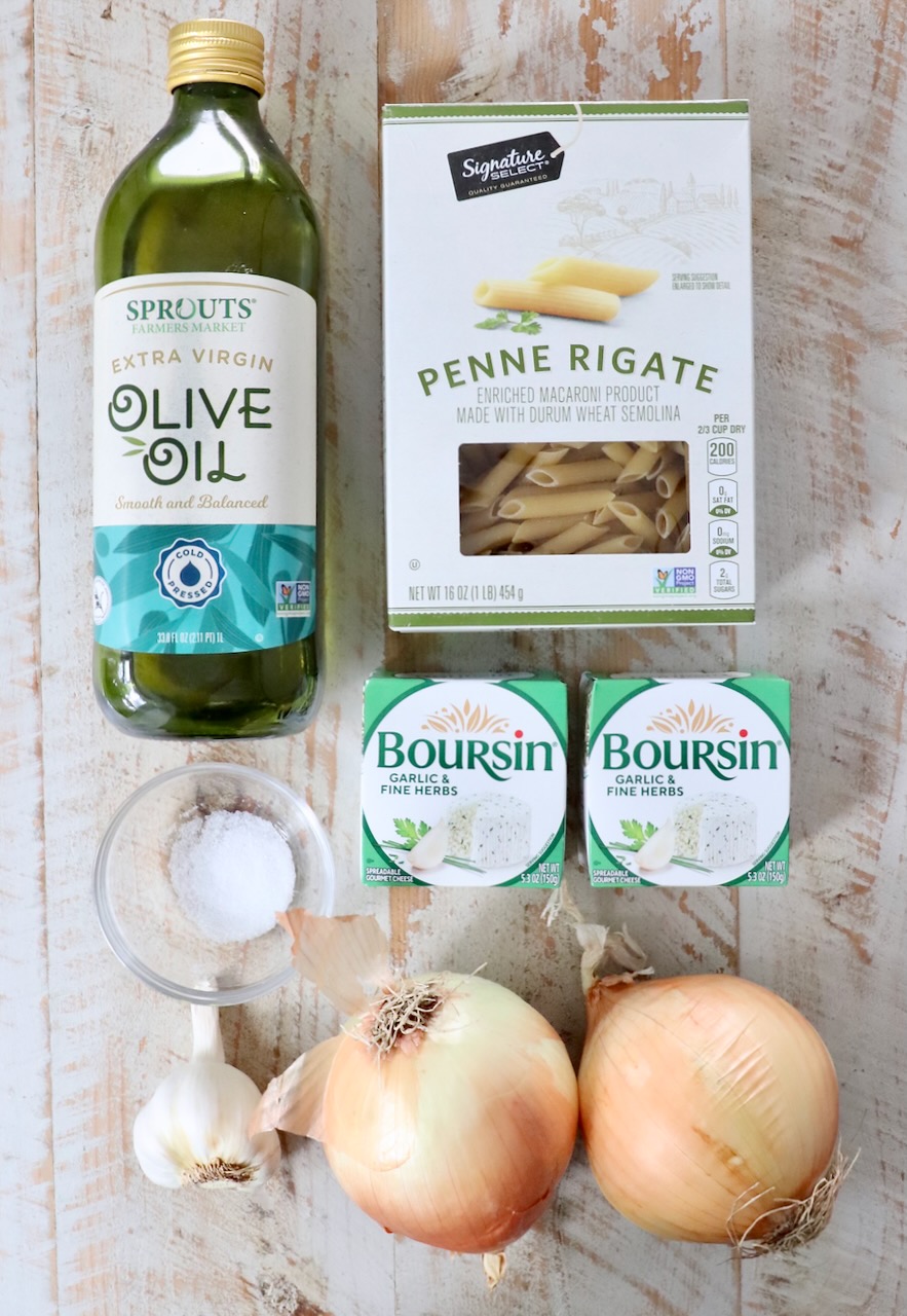 Ingredients on a wooden surface: olive oil, penne pasta, two packs of Boursin cheese, salt, garlic bulb, and two onions.