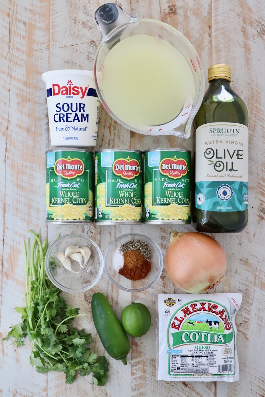 A flat lay of Mexican street corn soup ingredients, including sour cream, canned corn, olive oil, sour cream, spices, an onion, lime, jalapeño, cilantro, and cotija cheese on a wooden surface.