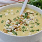 A creamy corn soup topped with crumbled cheese, cilantro, and golden corn kernels fills a white bowl.