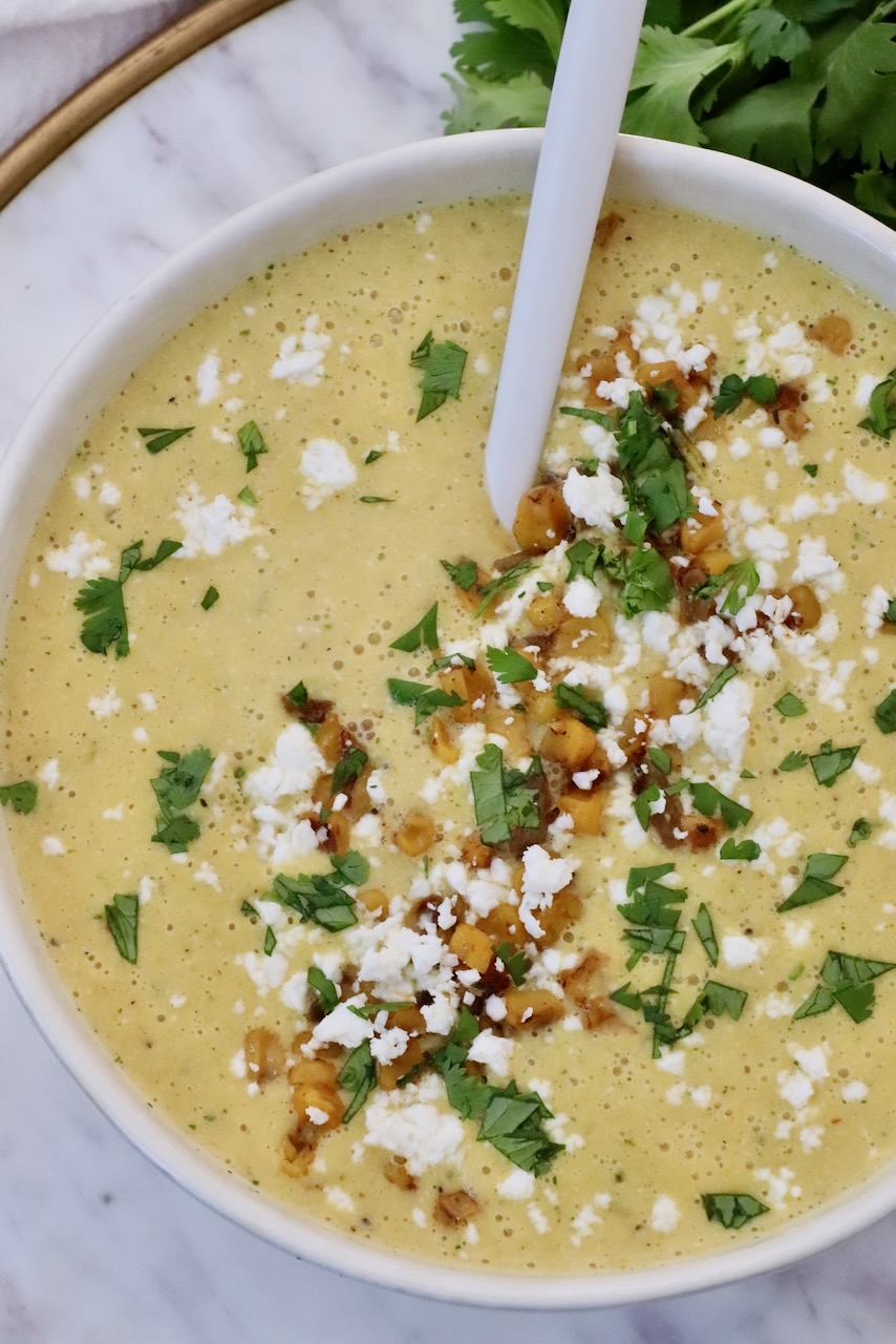 A creamy corn soup topped with crumbled cheese, roasted corn, and chopped green herbs, served in a white bowl with a spoon.