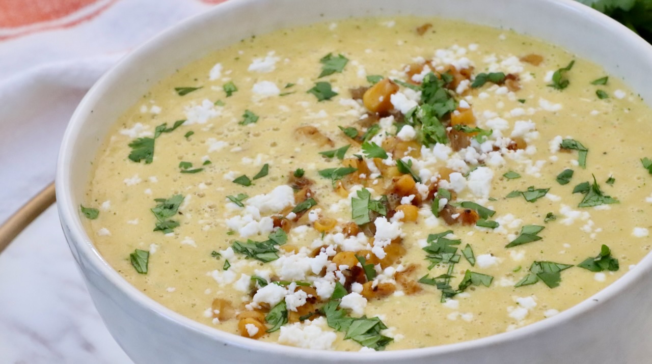 A creamy soup topped with crumbled cheese, cilantro, and golden corn kernels fills a white bowl. 