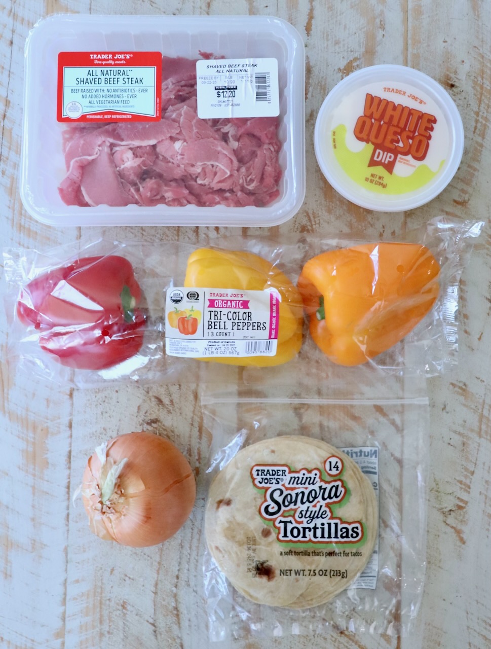 Assorted Trader Joe's ingredients on a wooden surface: shaved beef steak, white queso dip, tri-color bell peppers, a yellow onion, and mini tortillas.