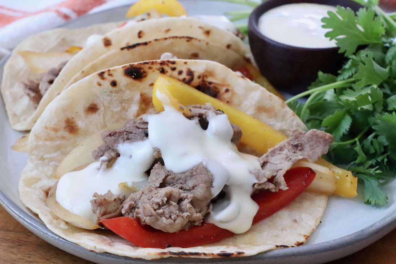 Grilled steak with yellow and red bell peppers, topped with creamy white queso in charred tortillas. Garnished with fresh cilantro on the side and a cup of queso on the side.