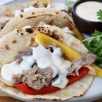 Grilled steak with yellow and red bell peppers, topped with creamy white queso in charred tortillas. Garnished with fresh cilantro on the side and a cup of queso on the side.