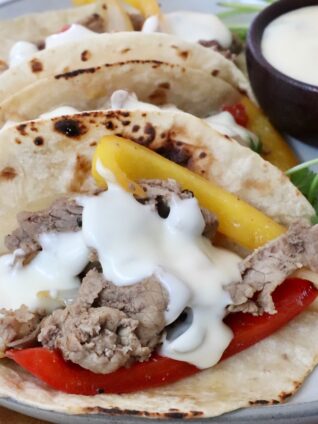 Grilled steak with yellow and red bell peppers, topped with creamy white queso in charred tortillas. Garnished with fresh cilantro on the side and a cup of queso on the side.