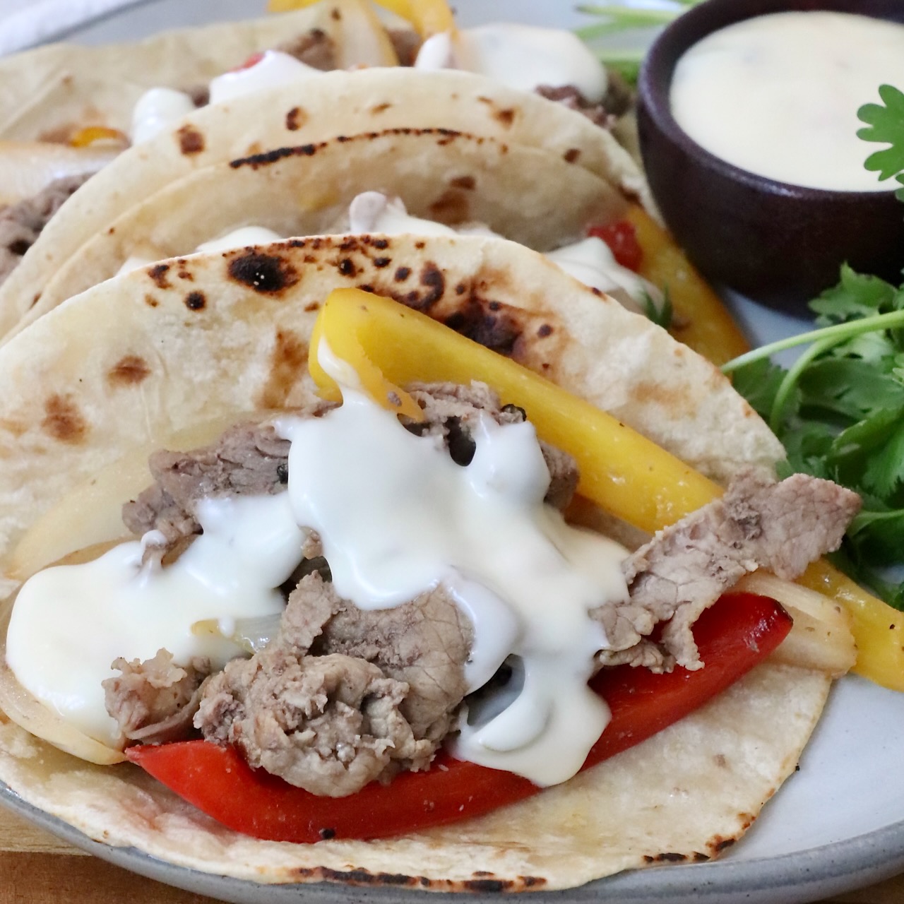 Grilled steak with yellow and red bell peppers, topped with creamy white queso in charred tortillas. Garnished with fresh cilantro on the side and a cup of queso on the side.