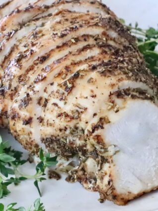 Sliced herb-crusted cooked turkey breast on a white plate, surrounded by fresh green herbs.