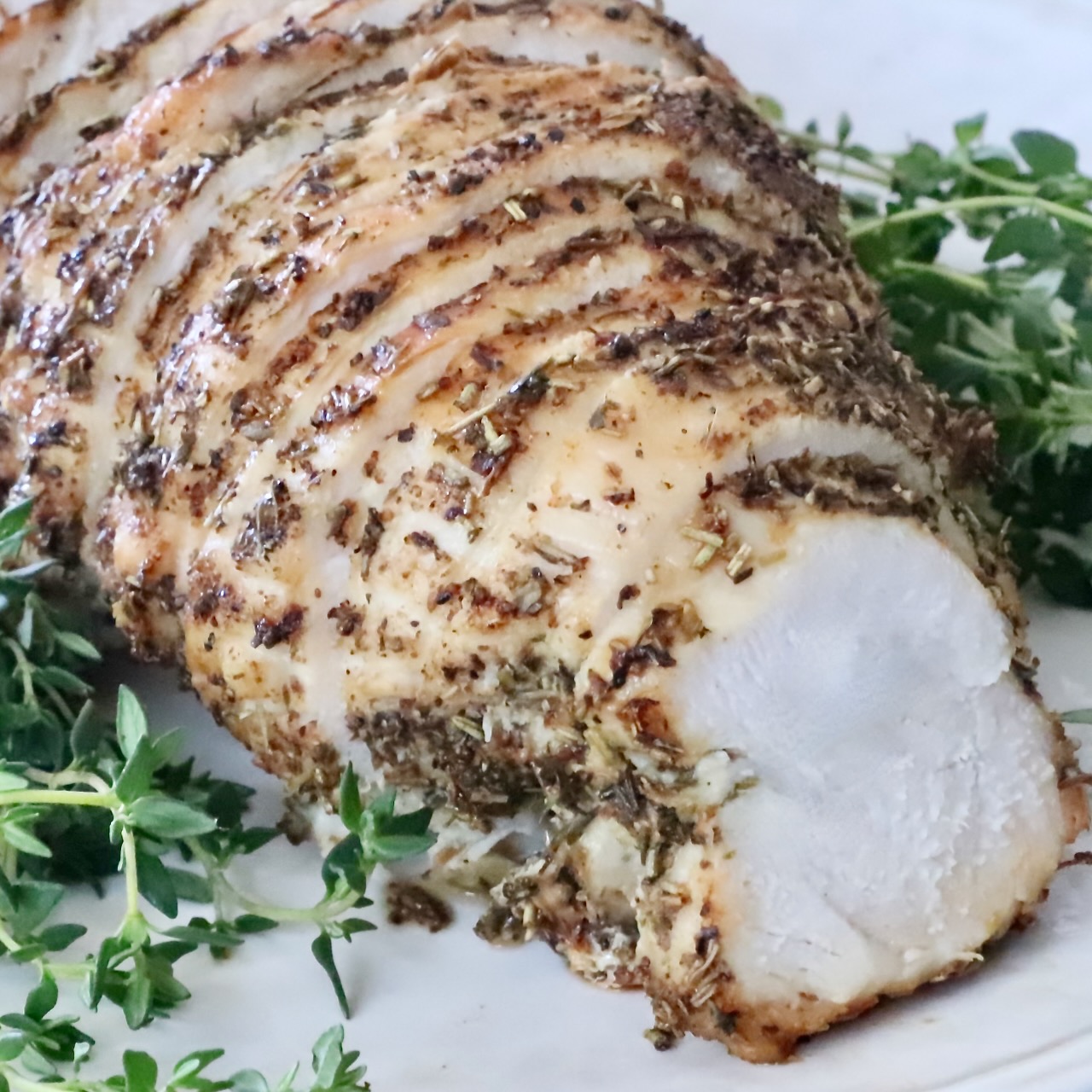 Sliced herb-crusted cooked turkey breast on a white plate, surrounded by fresh green herbs.