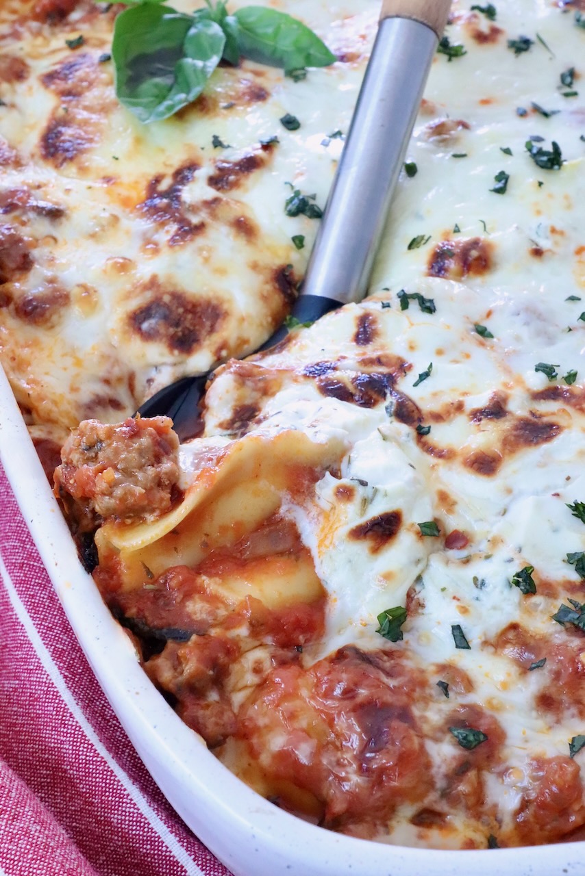 Close-up of a baked ravioli casserole in a white dish, topped with melted cheese and herbs. A serving spatula lifts a portion, showing layers of pasta and meat sauce.