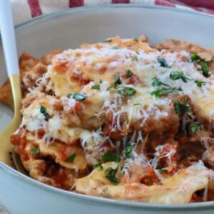 Close-up of a bowl of baked ravioli with melted cheese, garnished with fresh herbs with a fork in the bowl. Behind it, a red-striped cloth.