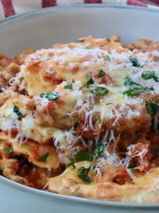 Close-up of a bowl of baked ravioli with melted cheese, garnished with fresh herbs with a fork in the bowl. Behind it, a red-striped cloth.