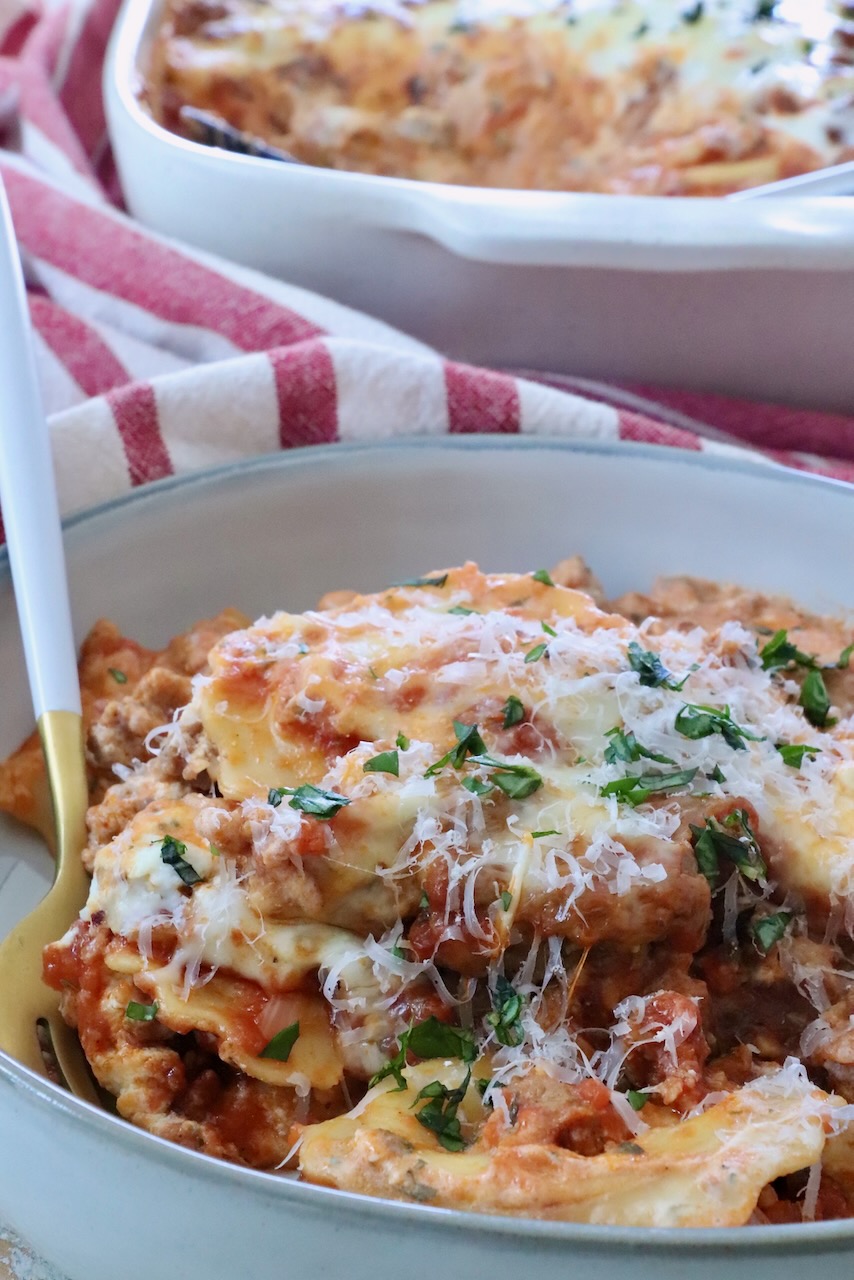 Close-up of a plate of baked ravioli with melted cheese, garnished with fresh herbs. Behind it, a red-striped cloth and a white casserole dish. 