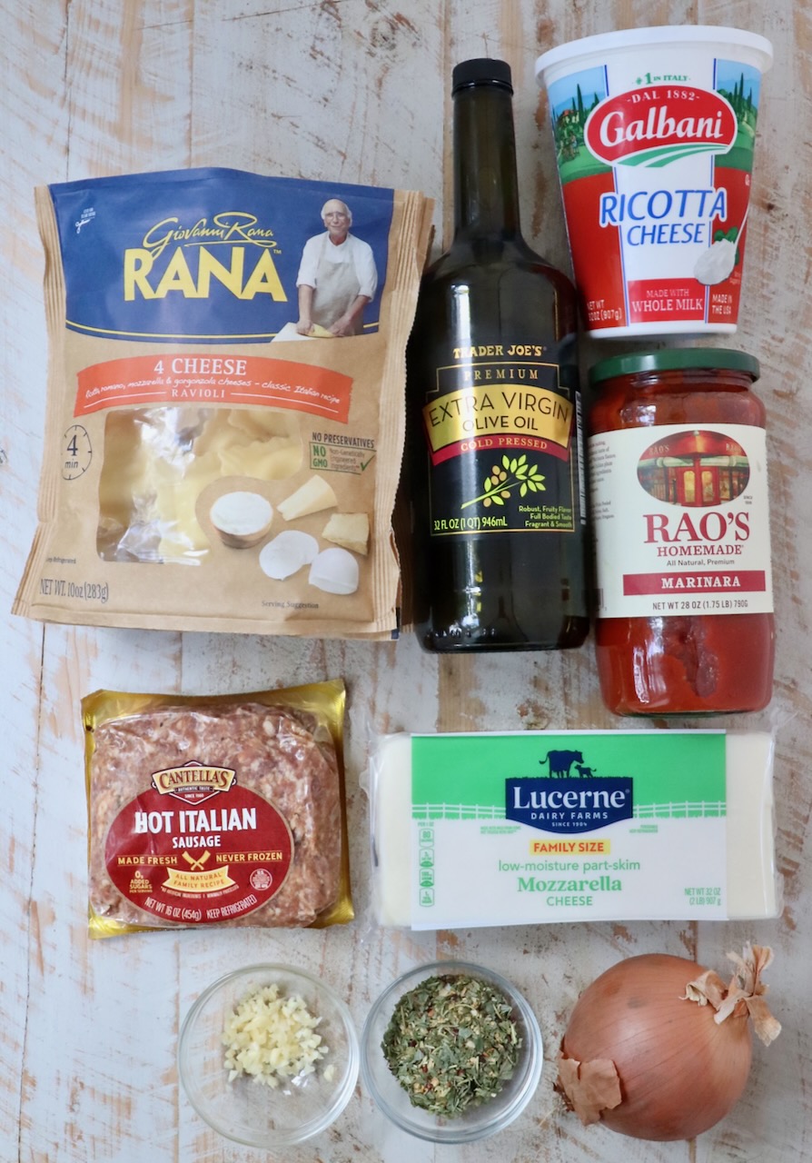 Assorted Italian ingredients on a wooden surface, including ravioli, olive oil, ricotta, marinara sauce, sausage, mozzarella, garlic, herbs, and an onion.