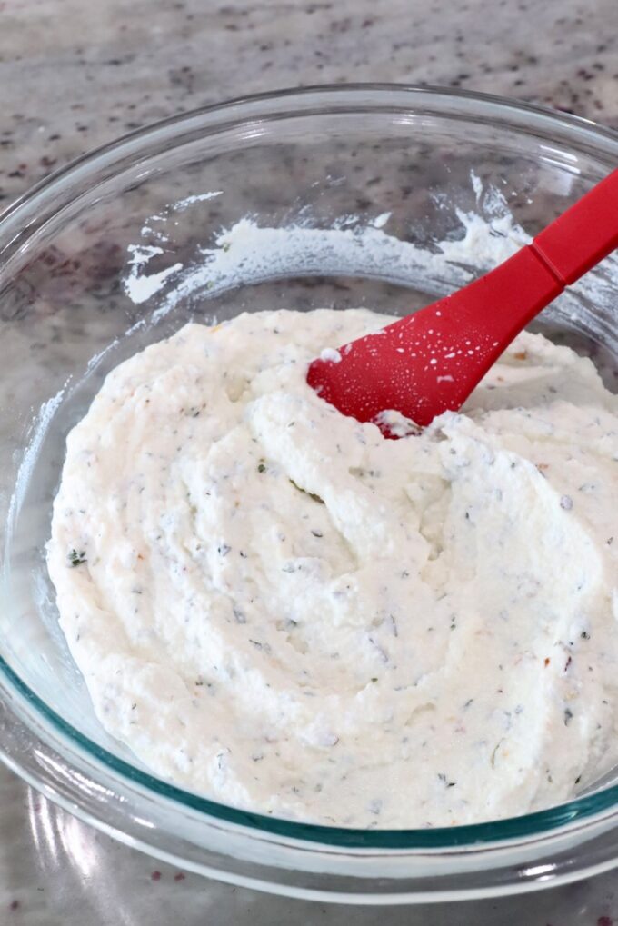 A glass bowl with fluffy, white ricotta cheese mixed with herbs. A red spatula is partially submerged, resting on a smooth marble countertop.