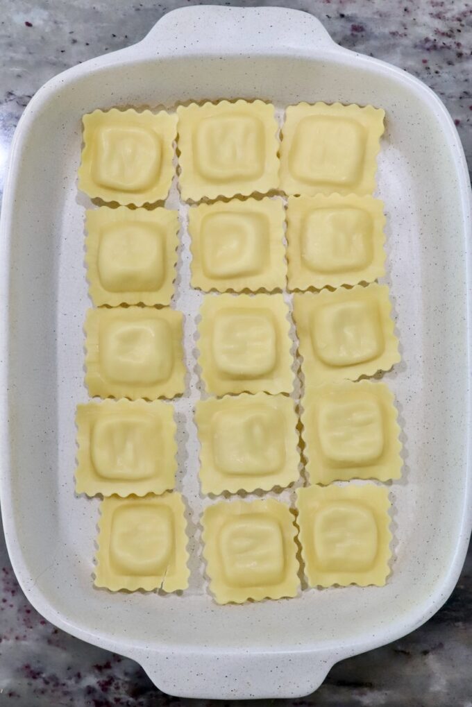 White baking dish with 15 uncooked ravioli in neat rows on a marble countertop. 