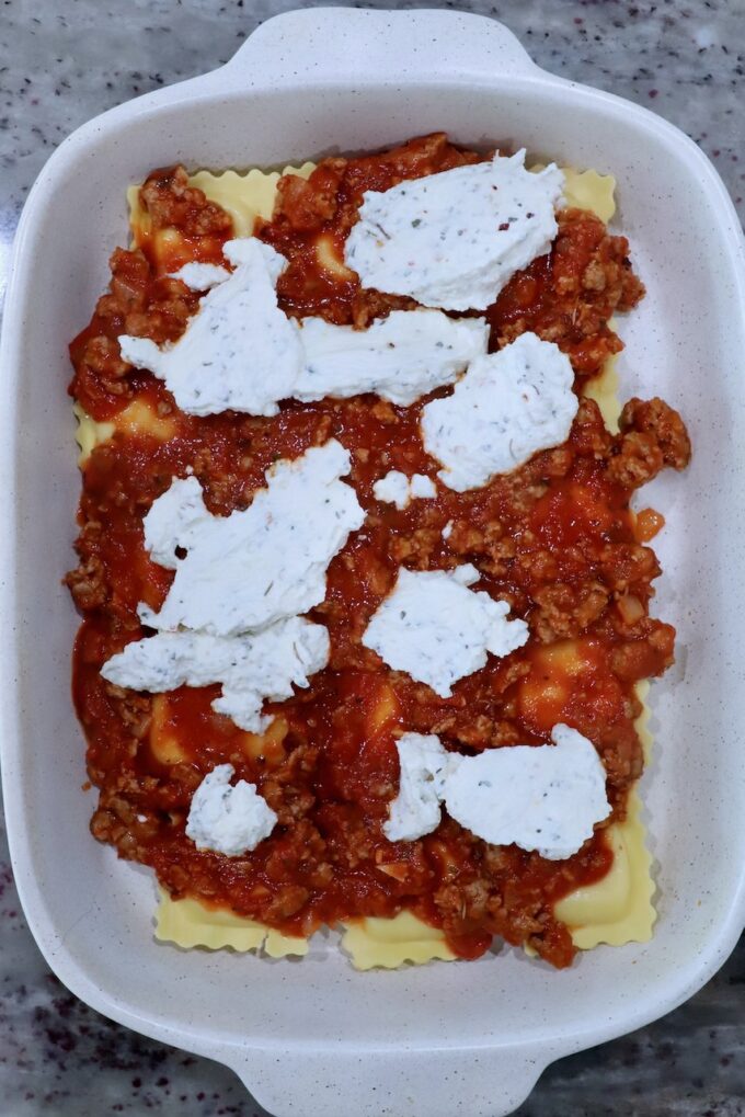 Baked ravioli preparation in a white dish on a speckled countertop. Layers of pasta are topped with meat sauce and dollops of ricotta cheese.