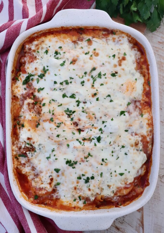 Cheesy baked ravioli topped with melted cheese and sprinkled with basil in a white baking dish. A red striped towel and fresh herbs are nearby.