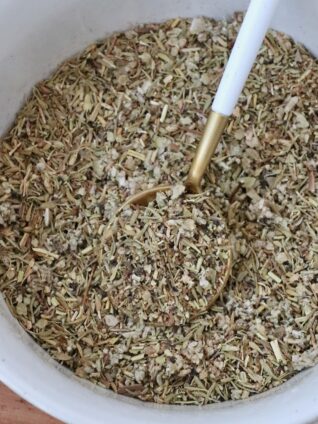 A bowl filled with a coarse, mixed herb blend featuring green and brown hues. A golden spoon with a white handle rests inside, evoking a rustic culinary vibe.