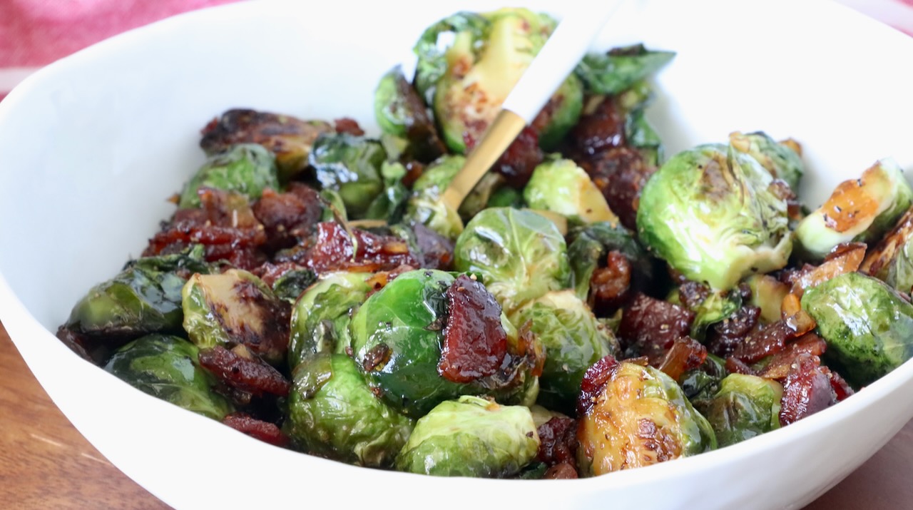 A white bowl filled with caramelized Brussels sprouts mixed with crispy bacon pieces, glistening with glaze, and a spoon for serving.